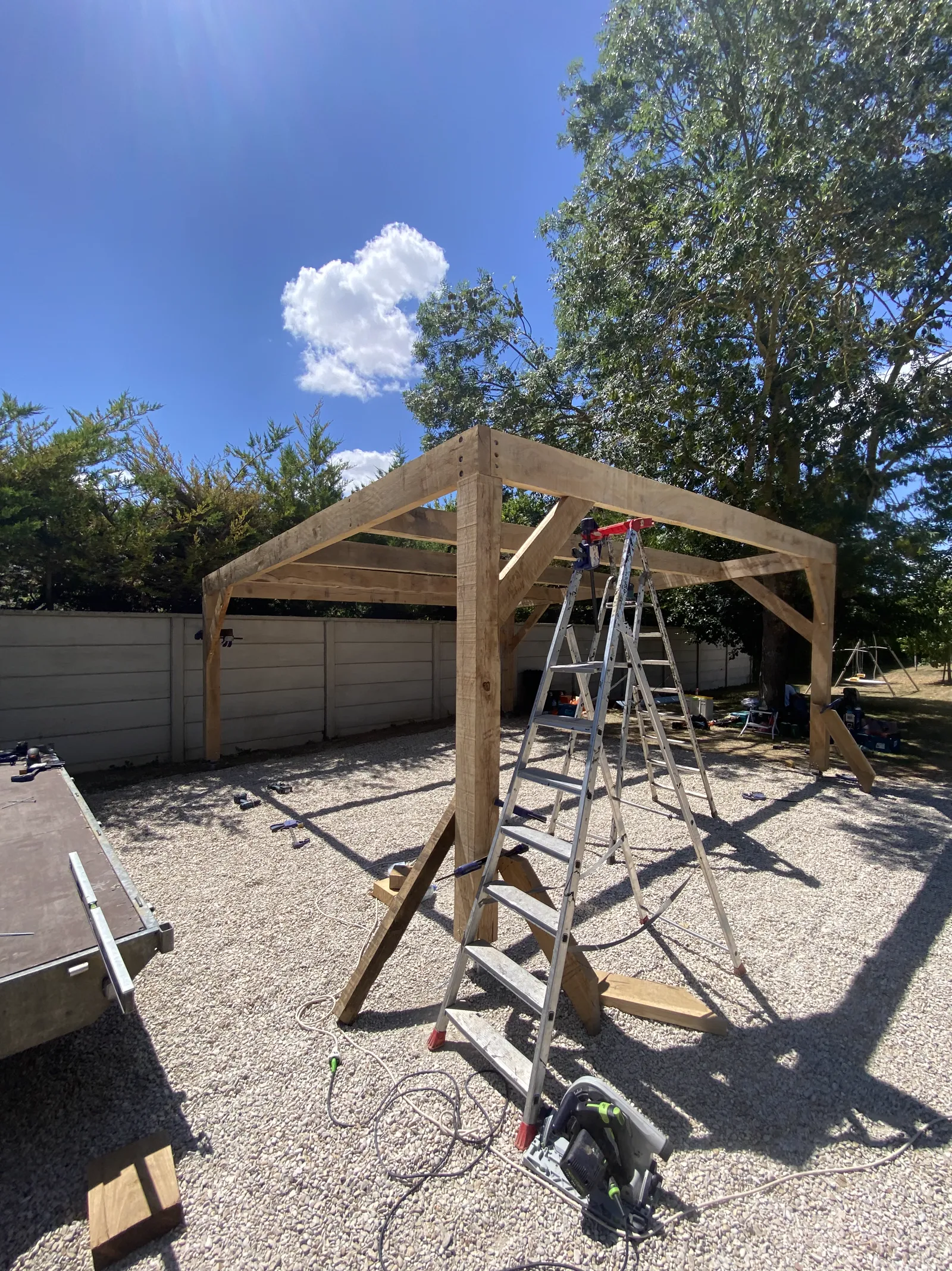 Charpente de carport en chêne massif brut de sciage en cours de montage à Pont-Sainte-Marie près de Troyes, jambes de force posées, escabeaux et outillage au sol sur un terrain en gravier, ciel bleu.
