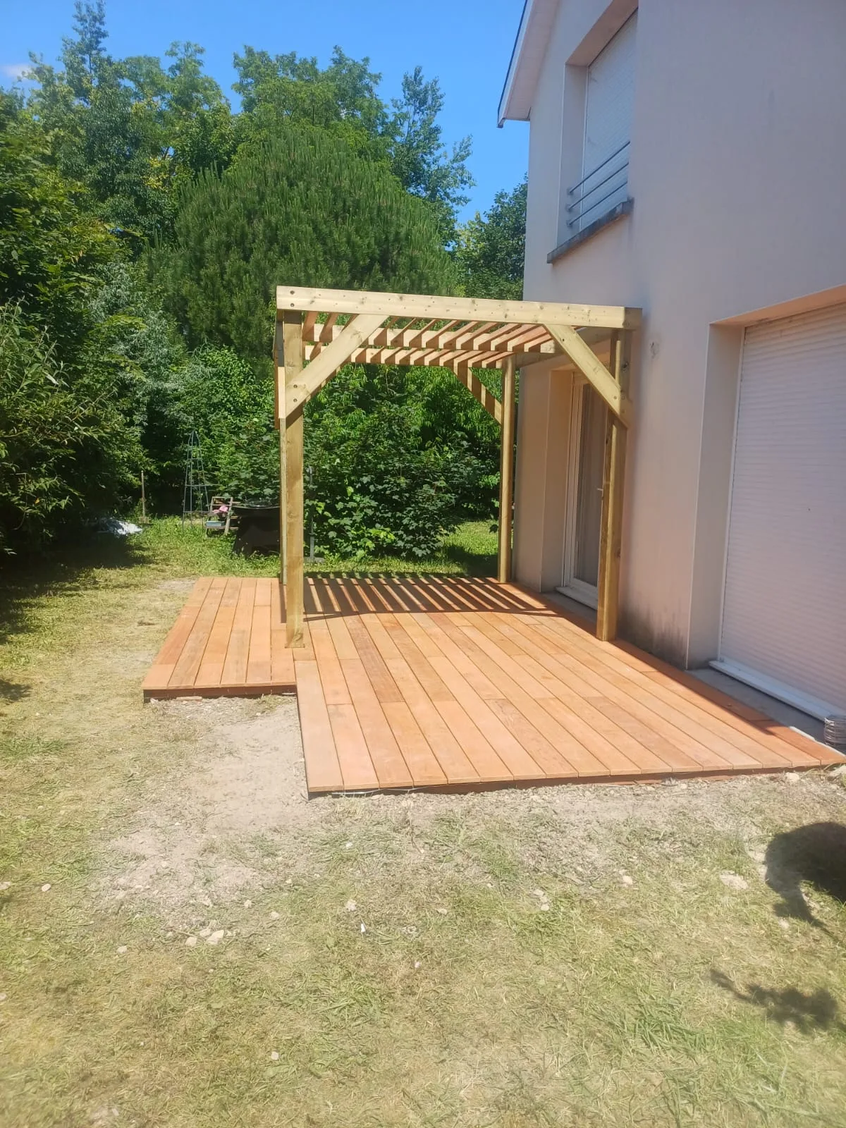 Terrasse en bois exotique avec pergola bois adossée vue depuis le jardin sous un ciel bleu, réalisée par La Planète Bois